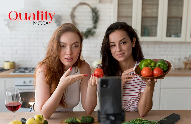 Dos mujeres en una cocina con vegetales en las manos, grabando una clase